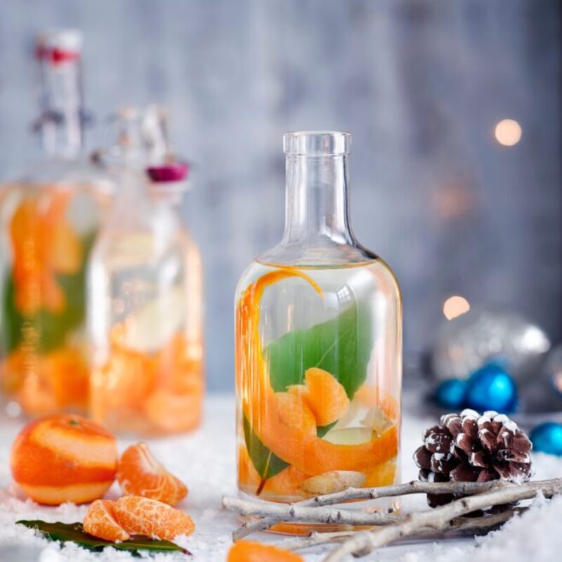 Rețetă de Crăciun Gin cu clementină, ghimbir și dafin pe o masă acoperită de zăpadă cu conuri de pin și segmente de clementină