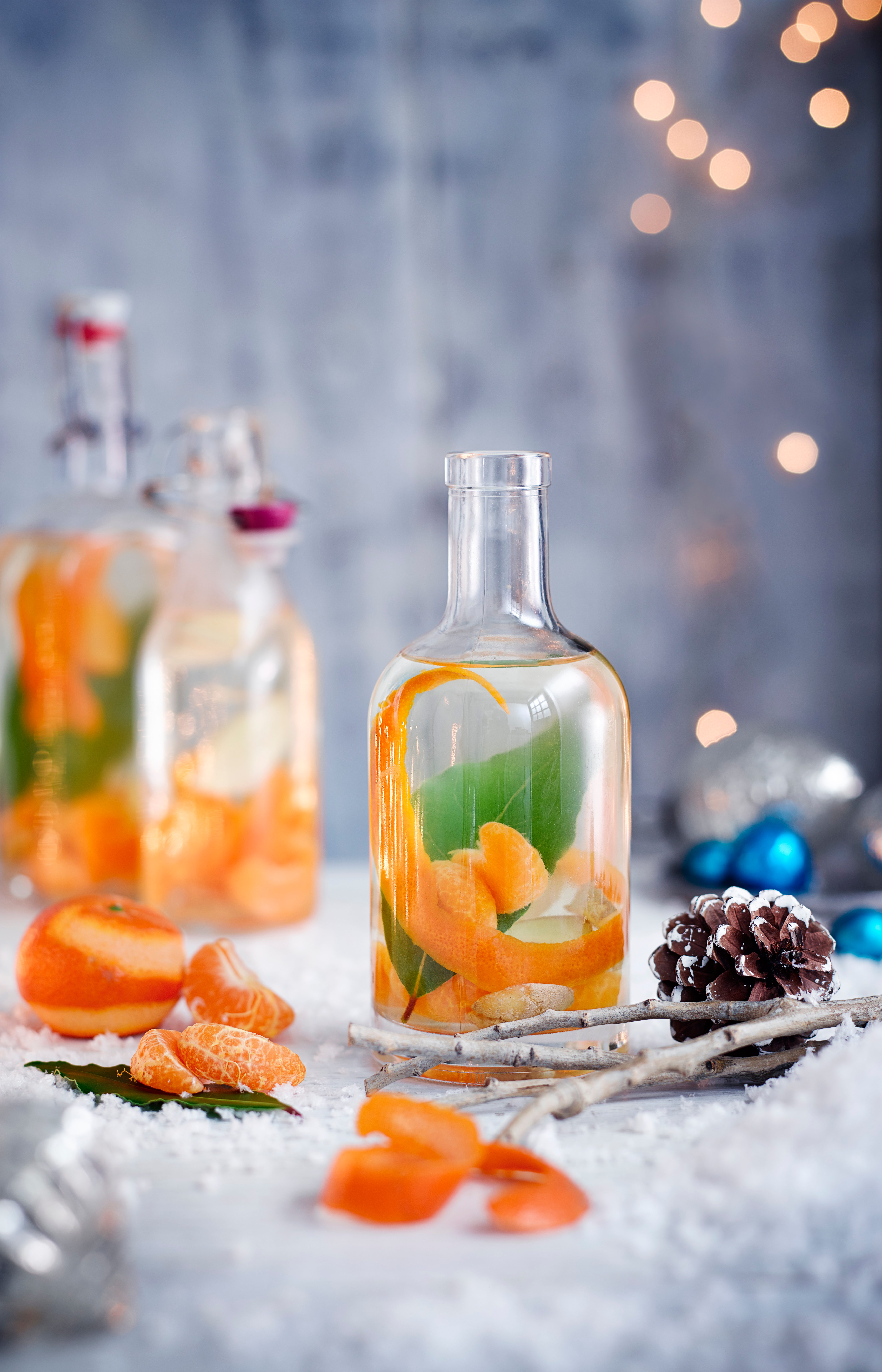Rețetă de Crăciun Gin cu clementină, ghimbir și dafin pe o masă acoperită de zăpadă cu conuri de pin și segmente de clementină