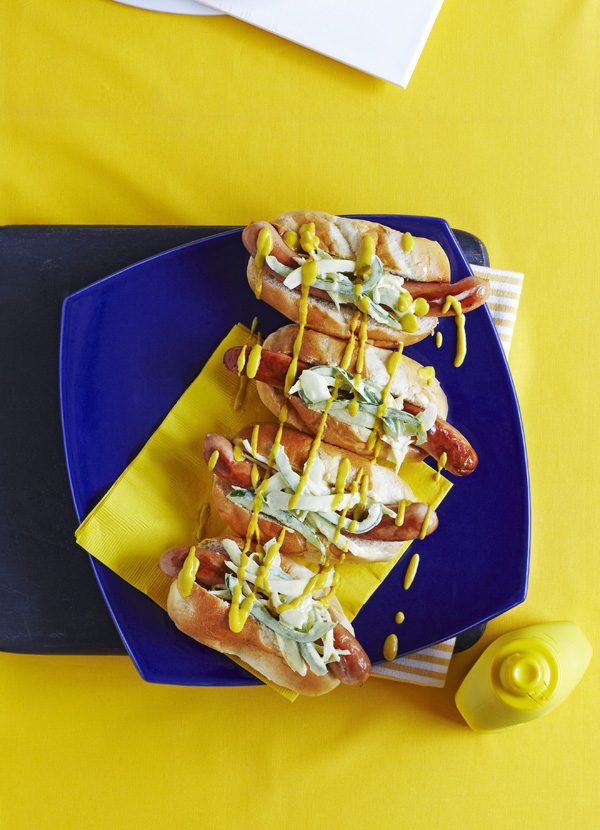O farfurie albastră acoperită cu patru chifle aurii pentru hot-dog, umplute cu cârnați lungi și răguță de varză