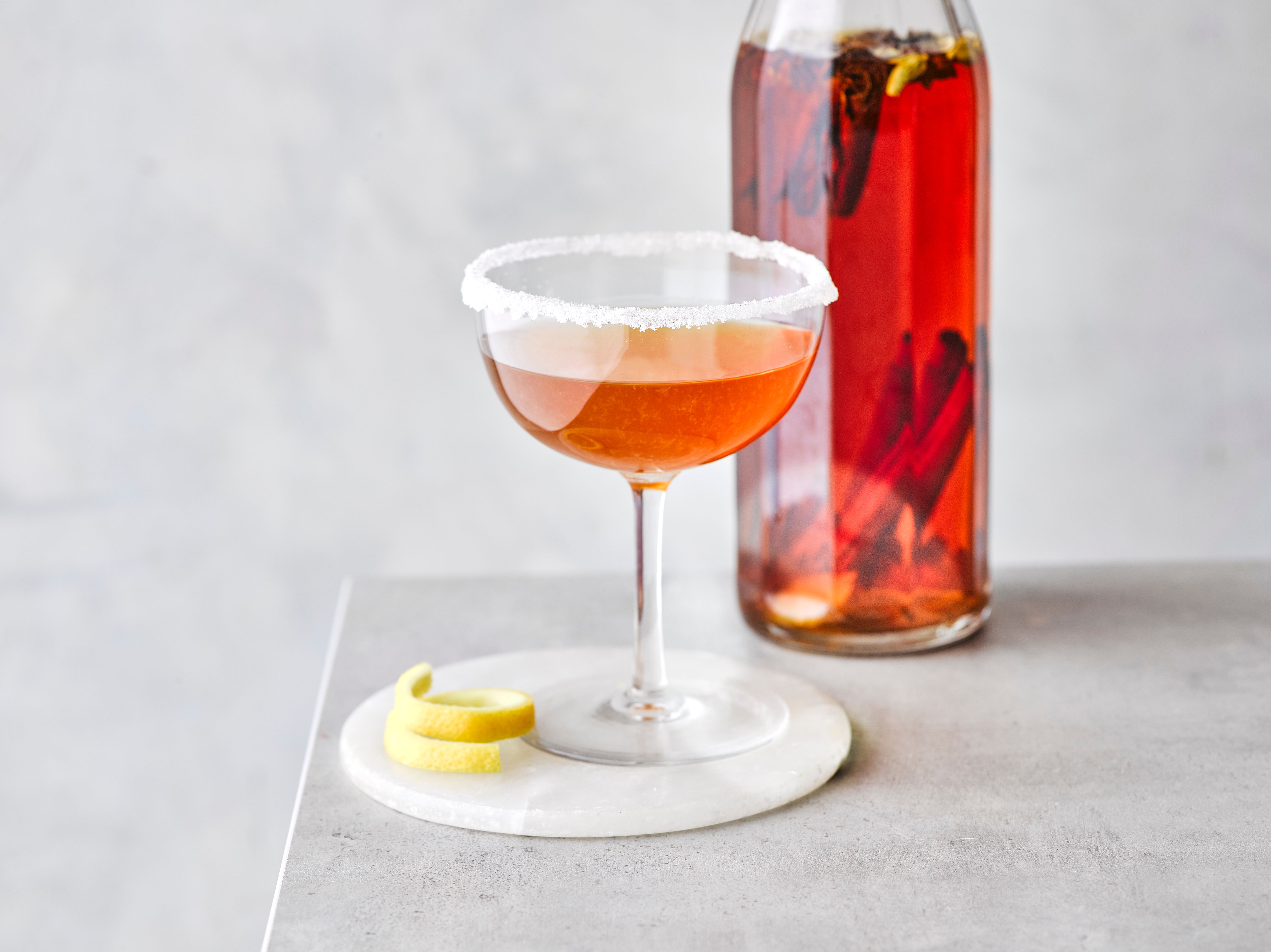 Cocktail sidecar într-un pahar coupe cu o sticlă de lichid roșu în spate